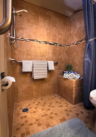 A tiled walk-in shower with a built-in bench holding rolled towels and a small plant. The shower has a grab bar with two towels hanging on it, a handheld showerhead, and a blue shower curtain partially visible on the right. The floor is tiled with small square tiles, and a blue bath mat is placed outside the shower area.