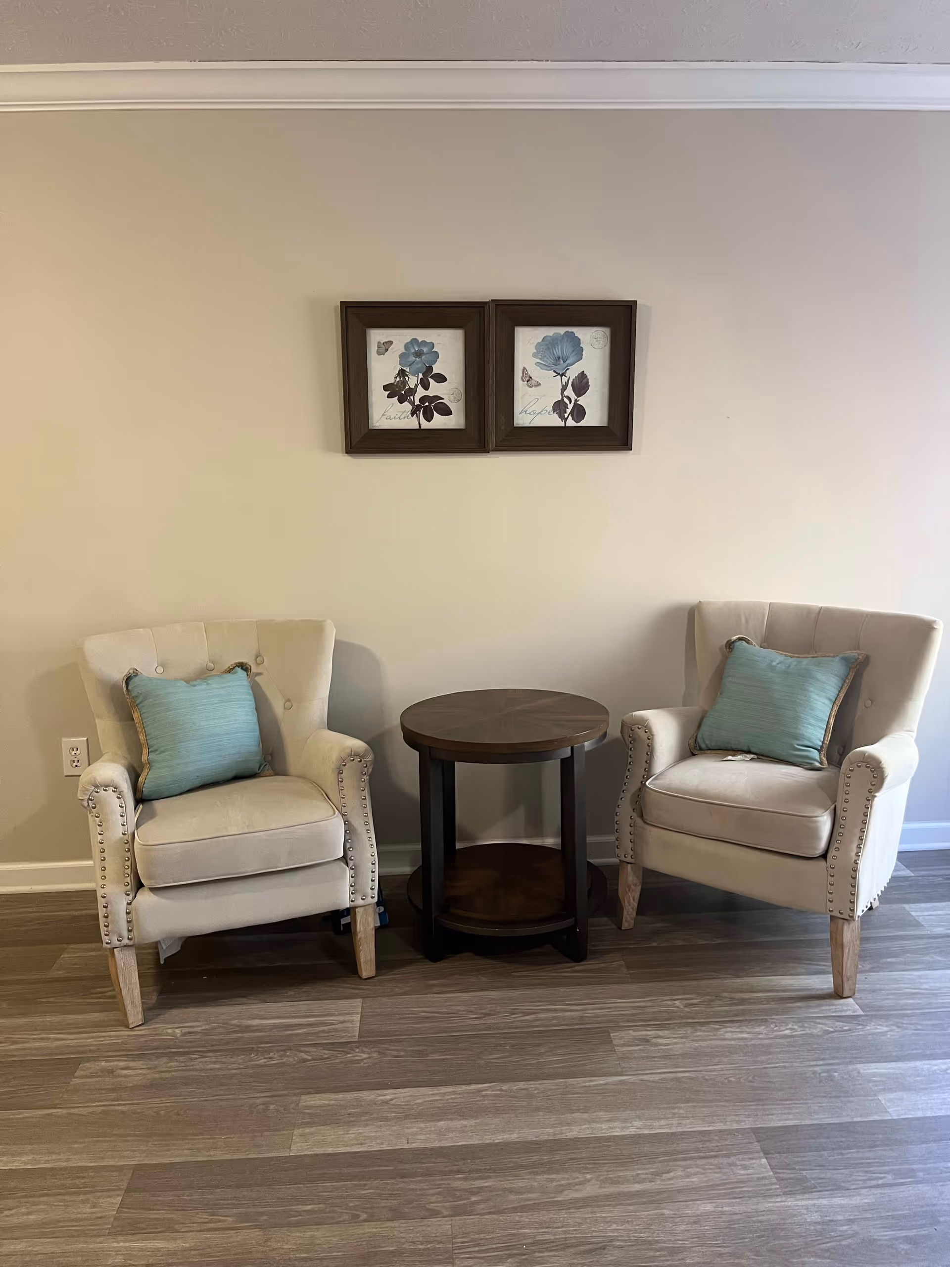 Two beige upholstered armchairs with light blue cushions are positioned on either side of a round wooden side table. Above the chairs, two framed floral artworks with blue flowers and butterflies hang on a beige wall. The floor is covered with wood-patterned vinyl or laminate.