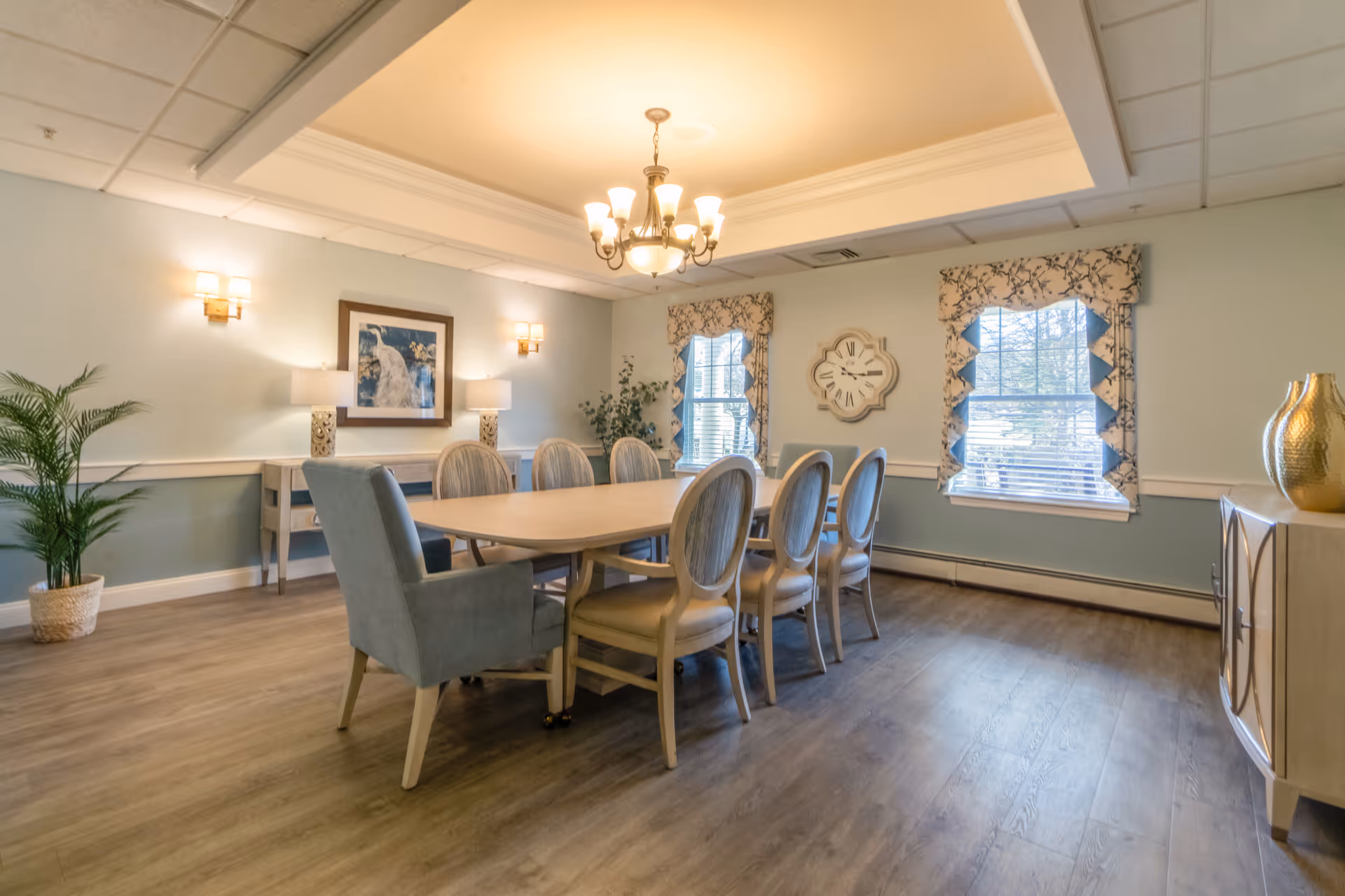 A well-lit dining room with a long wooden table surrounded by eight chairs, including two upholstered armchairs at the ends. The room features light blue walls, two windows with floral valances, a decorative wall clock, a framed picture, and a chandelier hanging from the ceiling. There are also two table lamps on a console table against the wall and a potted plant in the corner.