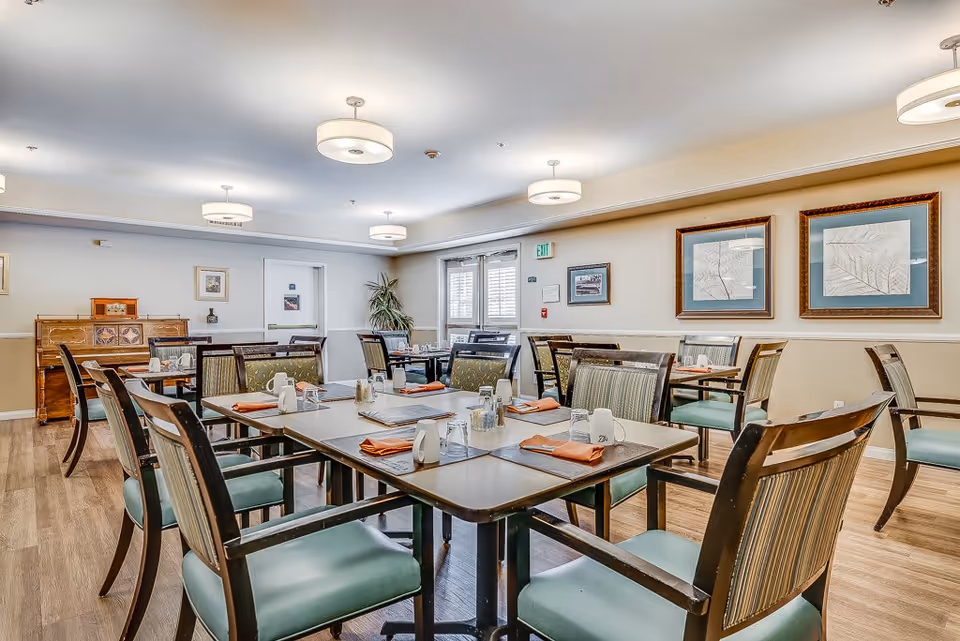 A well-lit dining room with multiple tables set with cups, glasses, and orange napkins. The room features wooden chairs with green cushions, framed artwork on the walls, and ceiling lights. There is a piano against one wall and a plant near a door with windows covered by white shutters.
