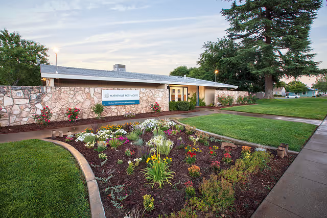 Exterior view of Marysville Post-Acute facility showing a single-story building with a stone facade, a sign with the facility's name, a landscaped flower bed with various colorful flowers, green grass, trees, and a sidewalk.