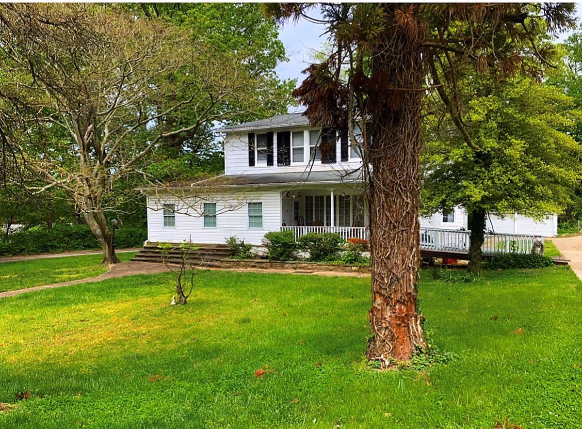 A two-story white house with black shutters surrounded by green grass and trees. There is a large tree trunk in the foreground and a pathway leading to the house. The house has a porch with white railings and steps leading up to it.