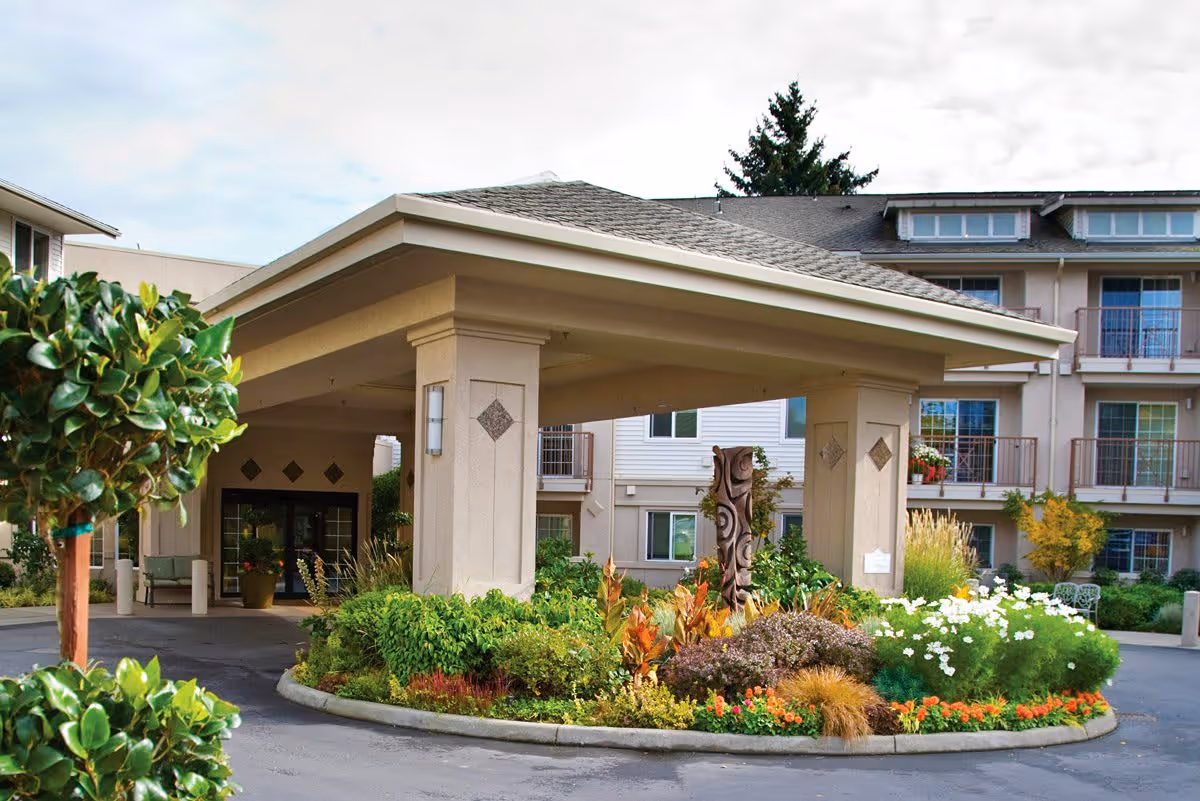 Entrance of The Lakeshore senior living facility with a covered drop-off area surrounded by well-maintained landscaping including various green shrubs, colorful flowers, and a decorative wooden sculpture. The building has multiple floors with balconies and large windows.