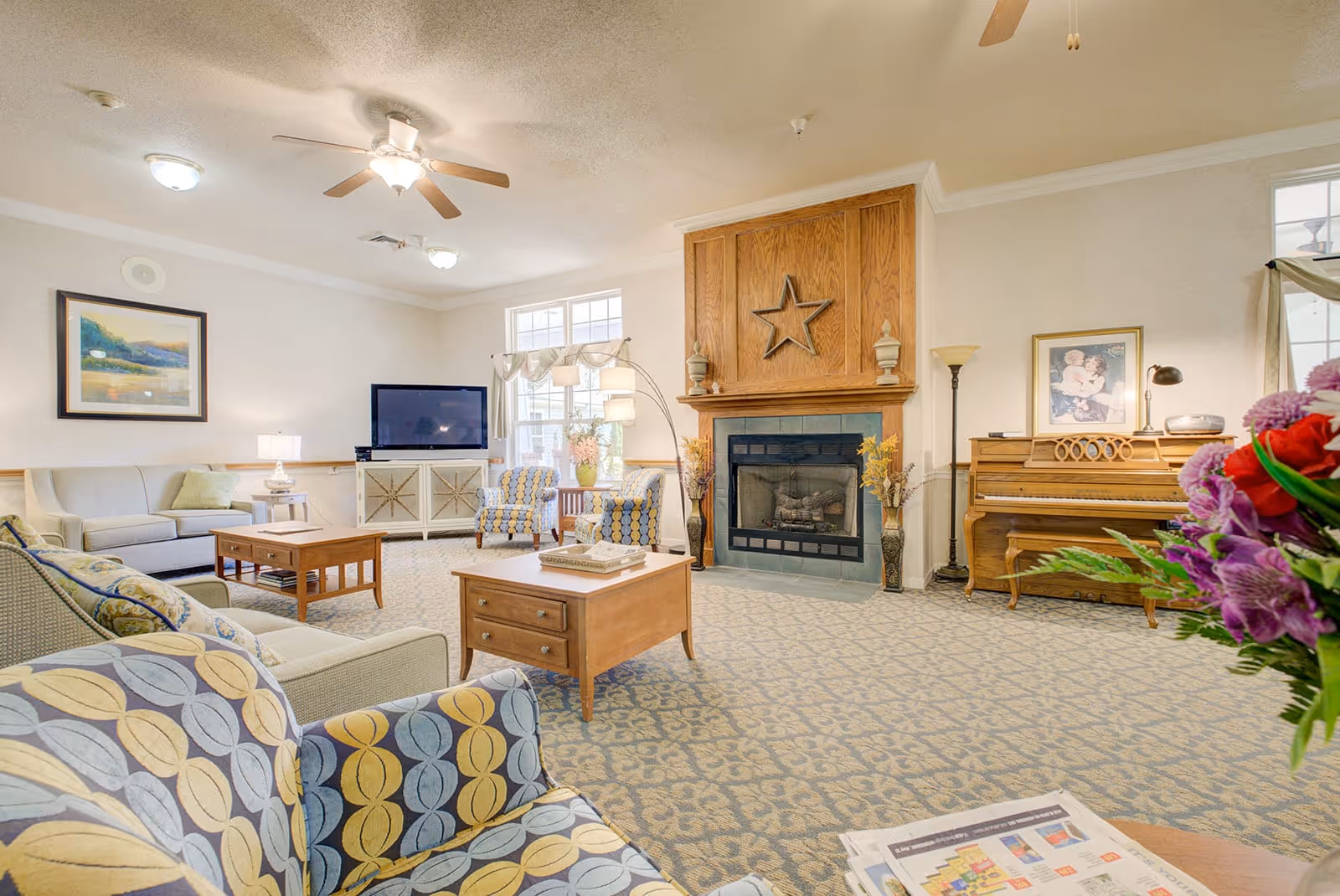A bright and spacious living room in an assisted living facility featuring patterned armchairs, a beige sofa, wooden coffee tables, a fireplace with a wooden mantle decorated with a star, a piano with a framed picture above it, a flat-screen TV on a white cabinet, and a vase of colorful flowers in the foreground.