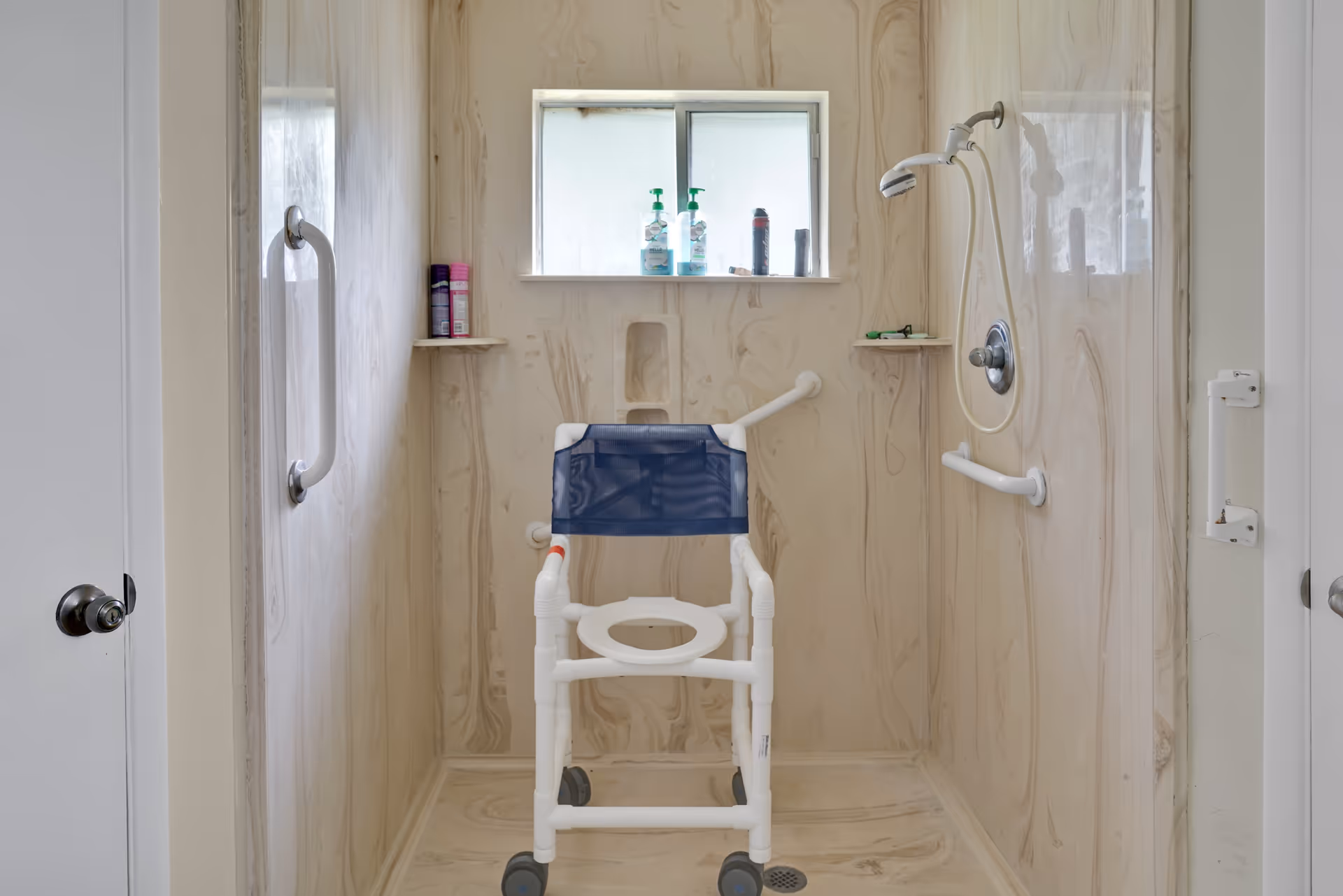 Accessible shower area with beige marbled walls, a window with toiletries on the sill, grab bars on the walls, a handheld showerhead, and a white shower chair with a blue backrest and wheels.