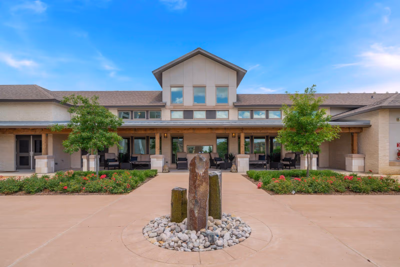 Front exterior view of a senior living facility with a central stone water feature surrounded by rocks, a paved walkway, landscaped flower beds with green shrubs and red flowers, two small trees, and a building with large windows and a covered porch with seating.