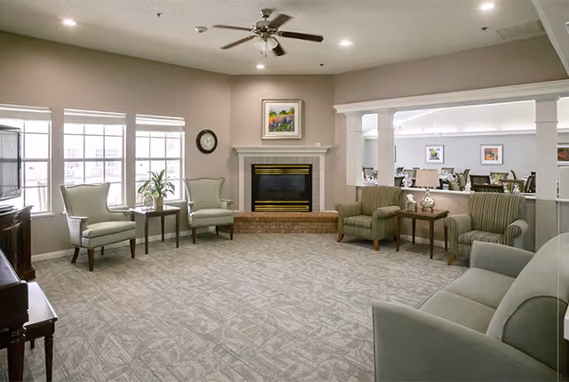 A spacious living room area with a beige carpet, light gray walls, and a ceiling fan. The room features a fireplace with a framed painting above it, two light green armchairs with a small table between them near the windows, and two striped armchairs with a small table between them on the right side. A gray sofa is partially visible in the foreground. The room opens into a dining area with tables and chairs visible through a large interior window.