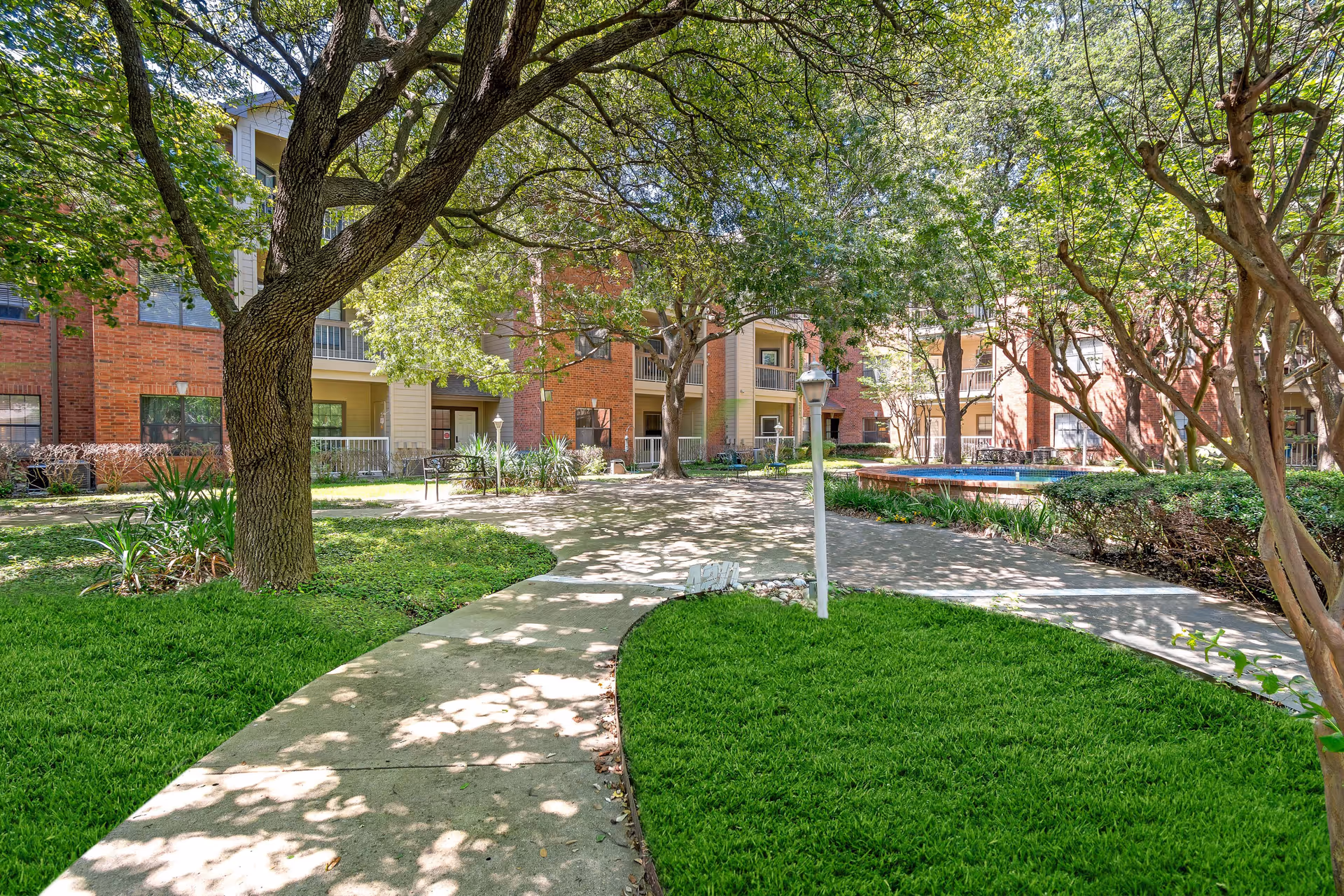 Outdoor courtyard area at Asher Point Independent Living of Sherman with a paved walkway, green grass, trees providing shade, and a multi-story brick and beige building in the background.