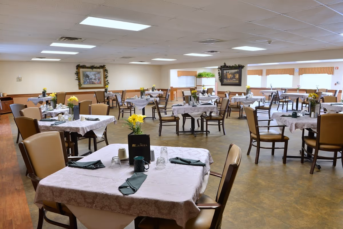 Spacious dining room with many tables covered in tablecloths, chairs, place settings and yellow flower centerpieces under fluorescent ceiling lights.