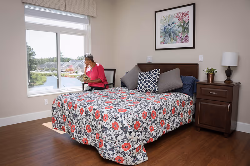A bedroom with a floral bedspread, wooden nightstand, and a person sitting by the window reading.