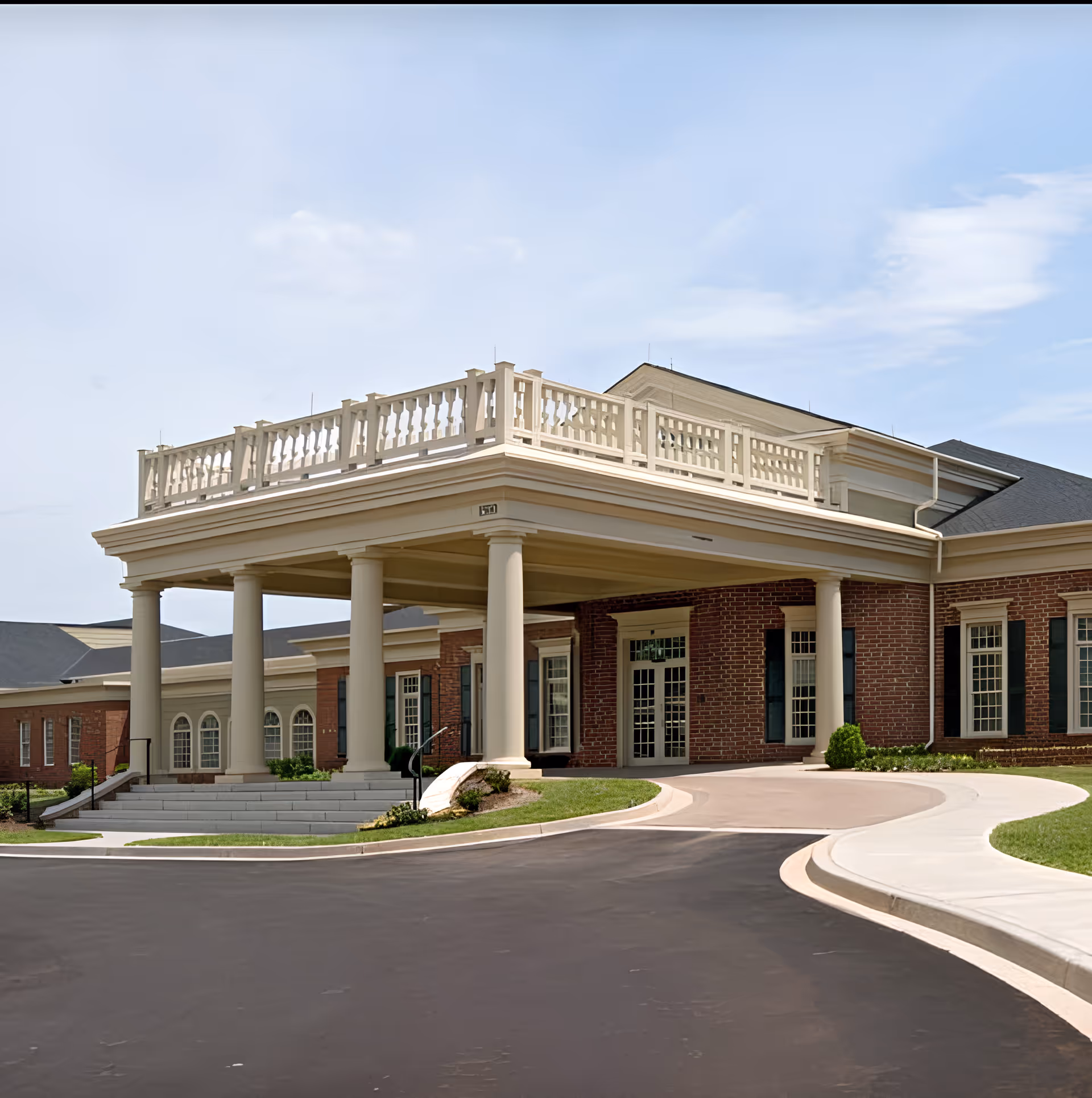 Front exterior view of The Woodlands at Furman facility showing a large covered entrance with white columns, brick walls, multiple windows, and a curved driveway.