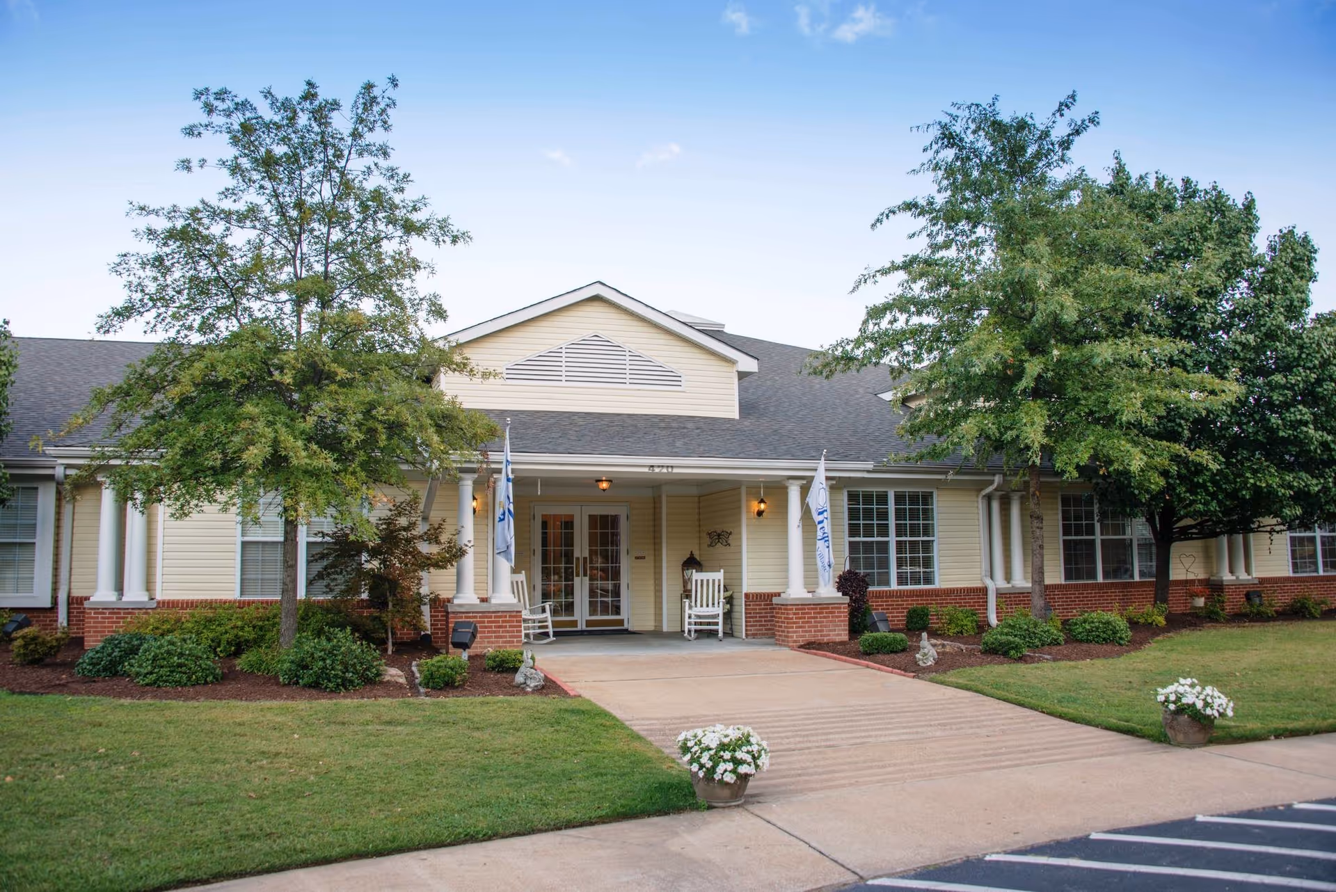 Front entrance of a single-story retirement facility with a covered porch, rocking chairs, trees, and landscaped lawn.