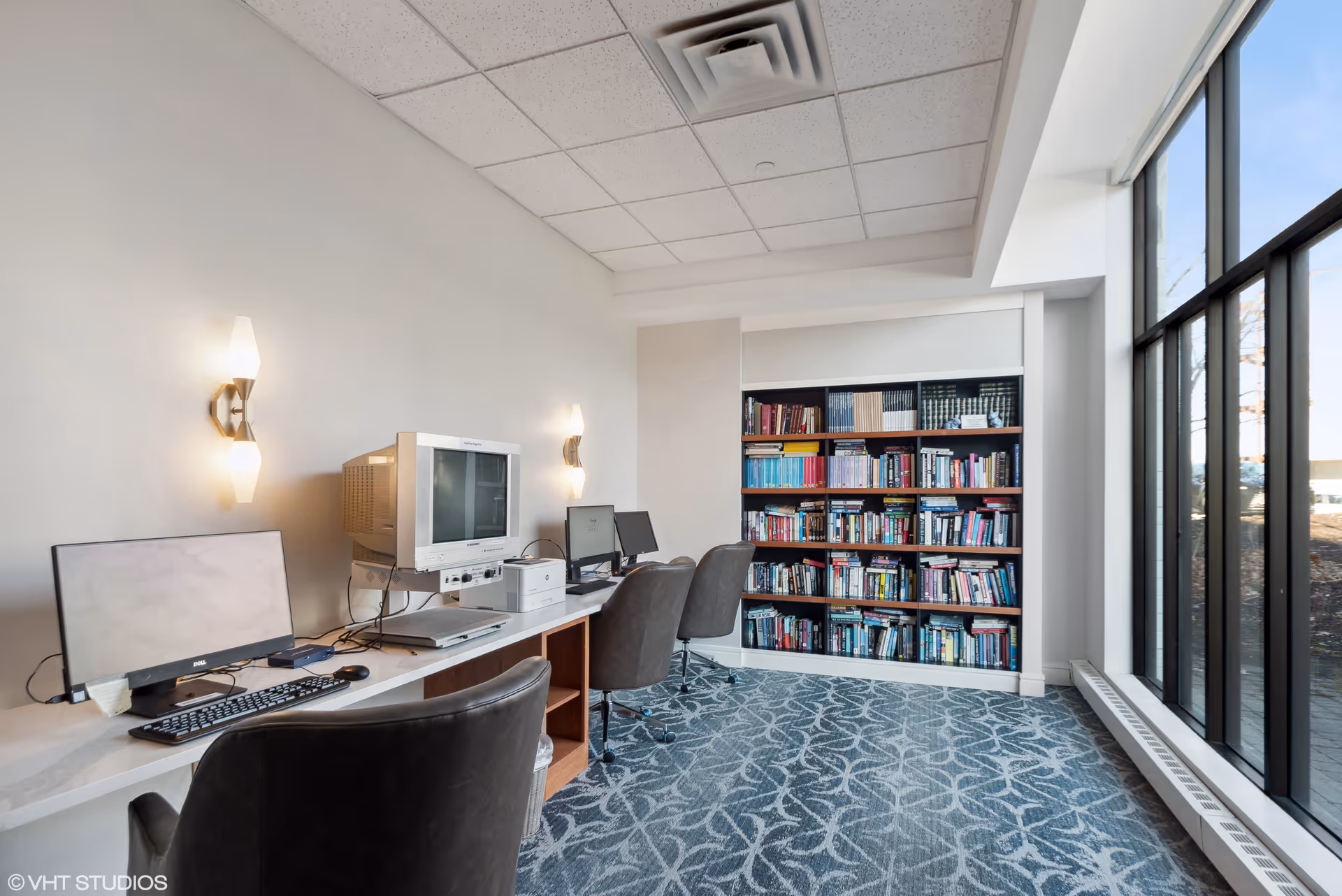 A bright room with a long white desk holding three computer stations, including an older CRT monitor and two flat screens. Three black office chairs are positioned in front of the desk. A large bookshelf filled with books is against the far wall. The room has patterned blue carpet and large windows letting in natural light.