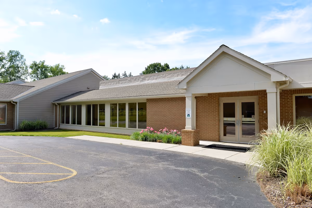 Exterior view of a single-story brick and siding building with a peaked entrance, large windows, and a paved parking area in front. There are some plants and flowers near the entrance under a clear blue sky.