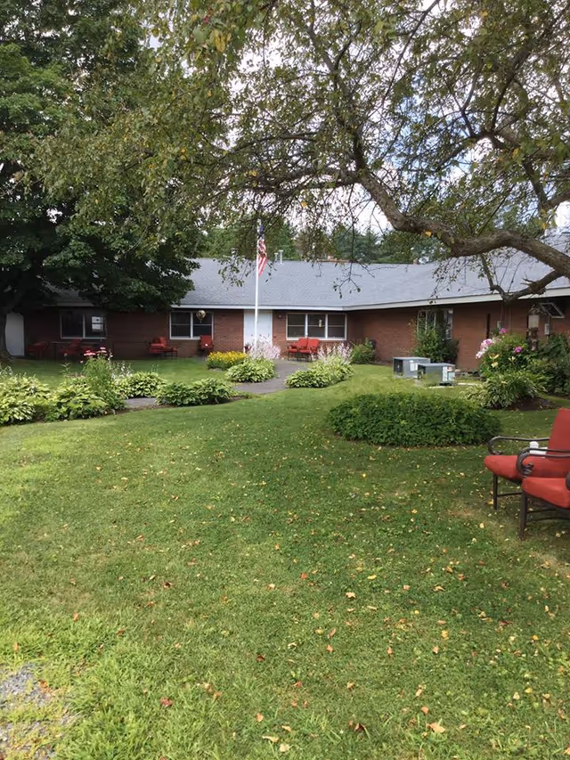 A single-story brick building with a gray roof surrounded by a well-maintained garden and lawn. There are several green bushes and flowering plants along a paved walkway leading to the building entrance. An American flag is mounted on a pole near the center of the building. Red cushioned chairs are placed outside near the windows and under a large tree with overhanging branches.