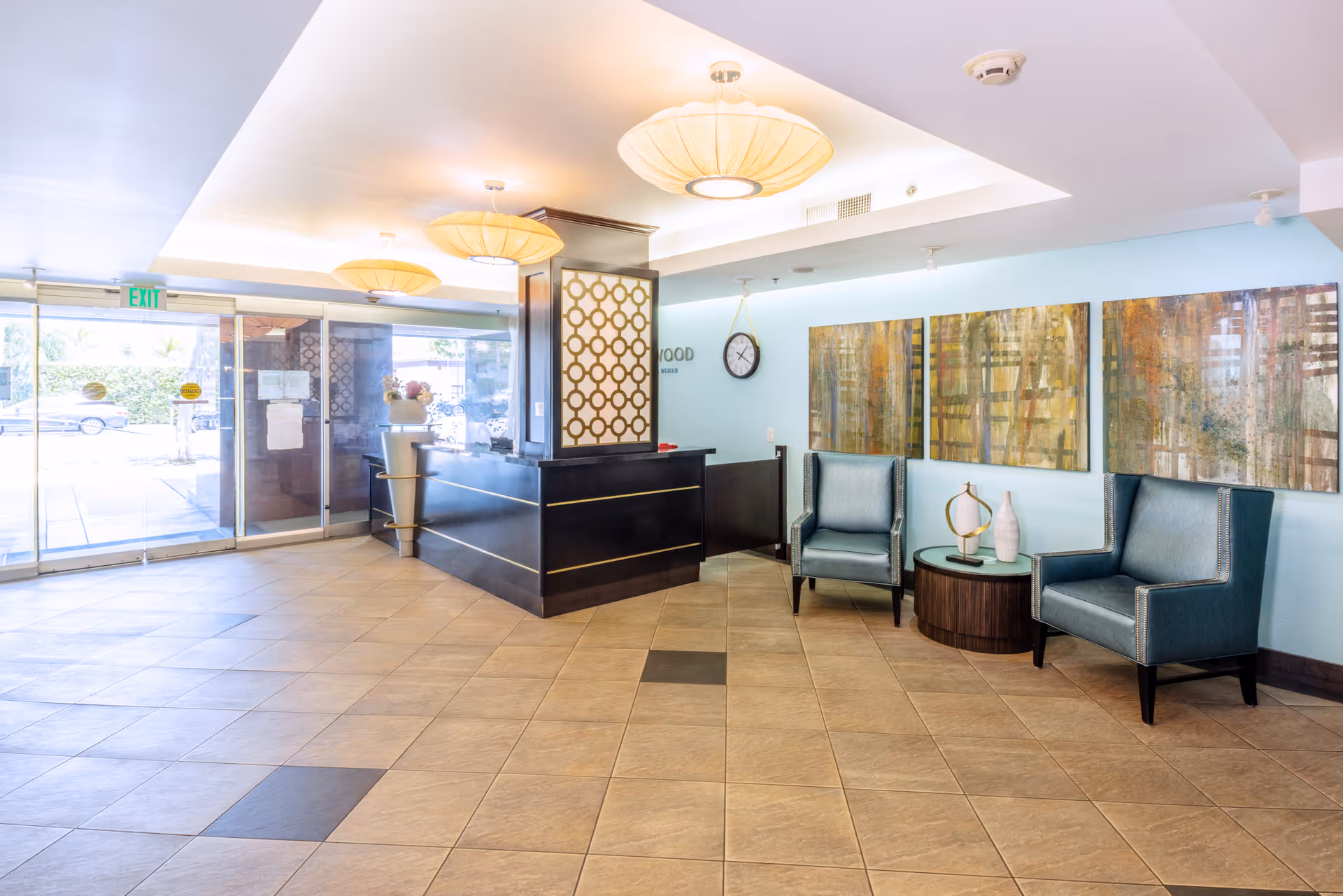 A bright and spacious reception area with a black front desk featuring gold accents. To the right, there are two blue upholstered armchairs with a round wooden table between them, decorated with modern vases and a sculpture. Behind the chairs, three abstract paintings hang on a light blue wall. The floor is tiled in a beige and brown pattern, and large glass doors lead outside. The ceiling has recessed lighting and three large, round pendant lights.