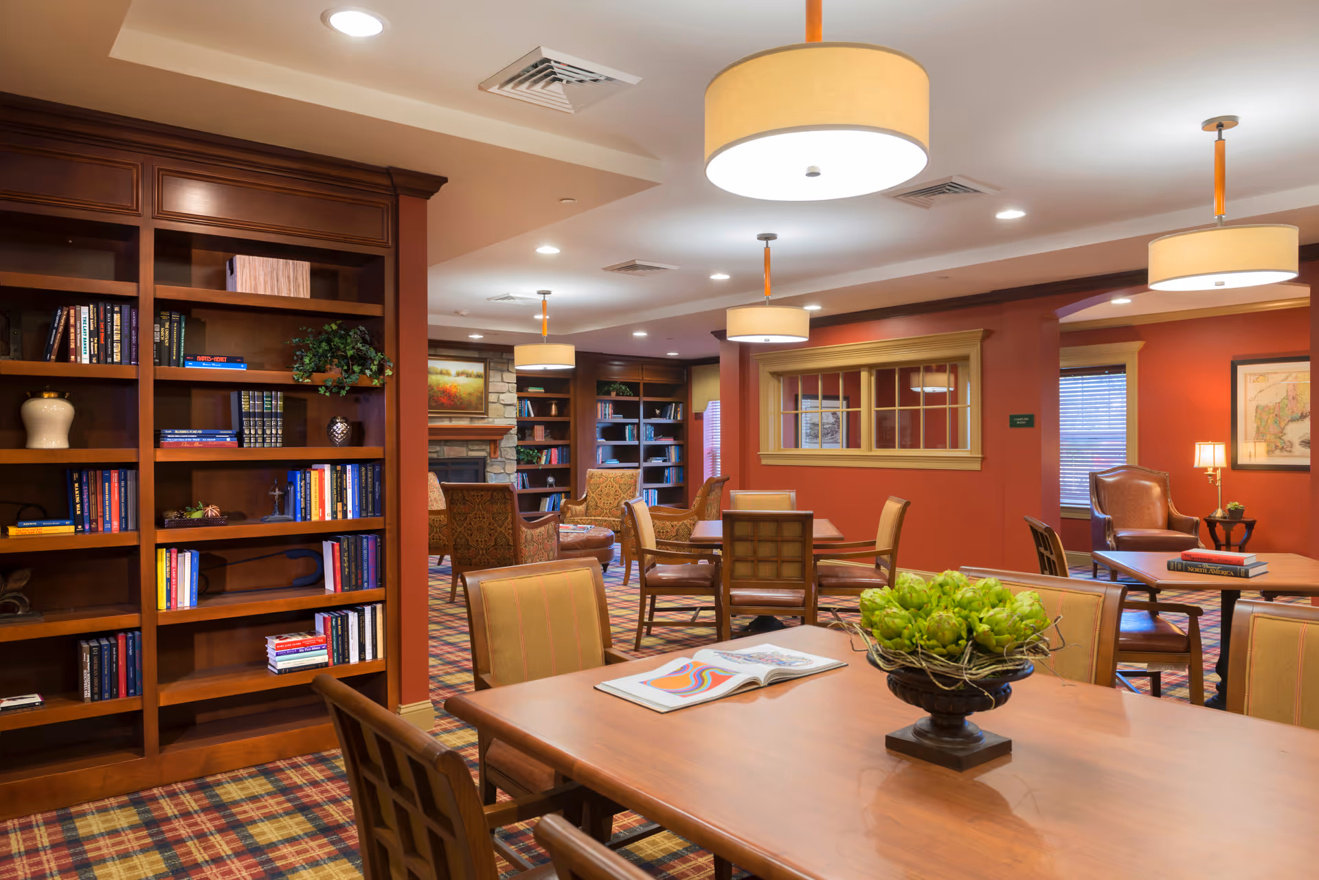 A warm communal library-style lounge with bookshelves, tables and chairs, pendant lights, and a fireplace in the background.