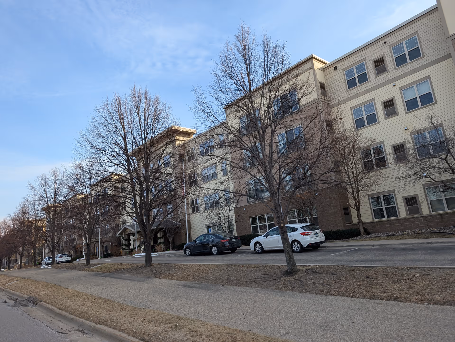 Exterior view of a multi-story senior living facility building with several leafless trees and parked cars along the driveway under a clear blue sky.