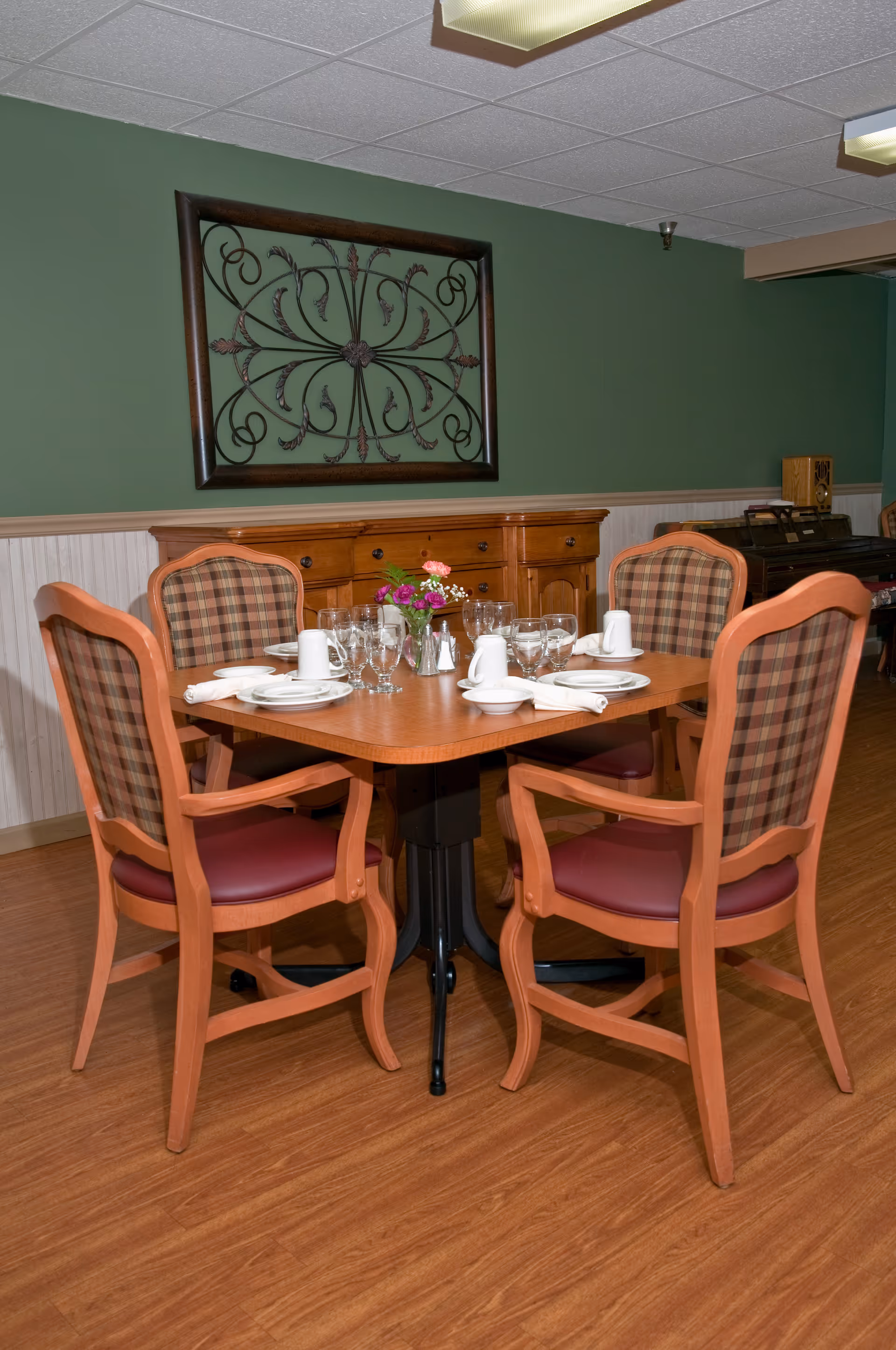 A dining table set for four with plates, glasses, napkins, and a small flower arrangement in the center. The table is surrounded by four wooden chairs with plaid upholstered backs and red cushioned seats. Behind the table is a wooden sideboard and a decorative wrought iron wall hanging on a green wall.