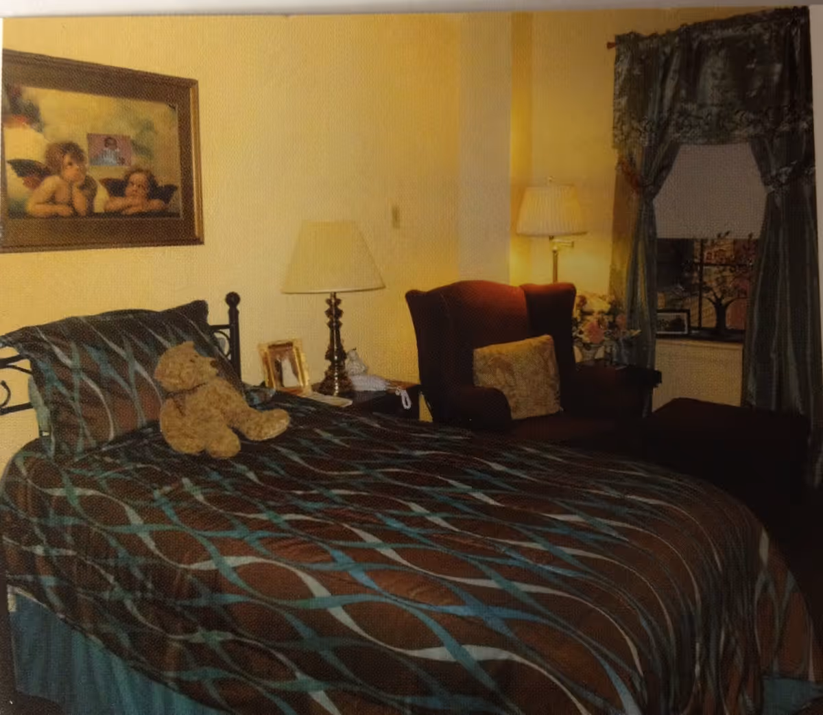 A cozy bedroom with a bed covered in a brown and blue patterned bedspread, a teddy bear sitting on the bed, a nightstand with a lamp and picture frames, a burgundy armchair with a cushion, and a window with decorative curtains.