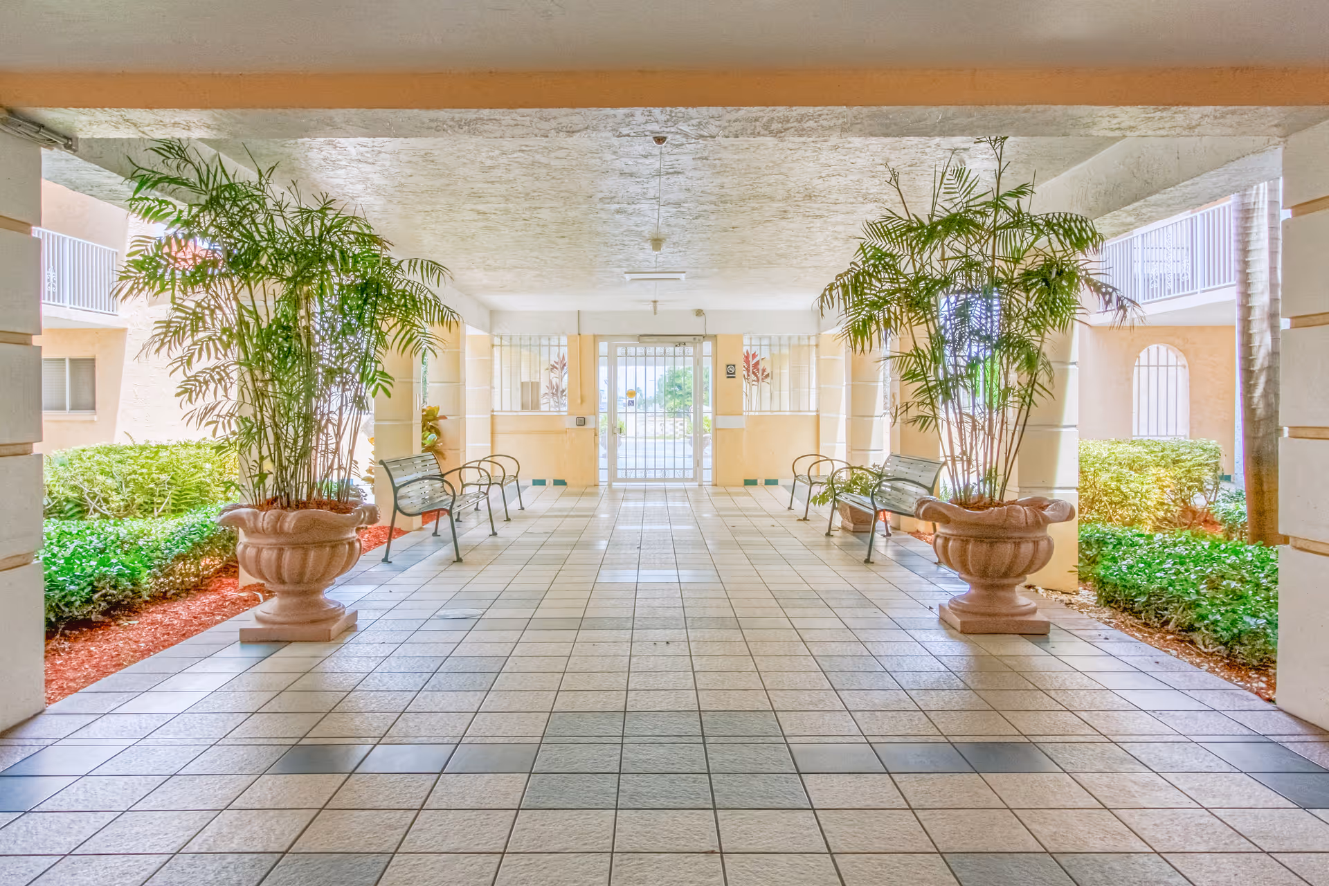 Covered outdoor walkway with tiled floor, large potted plants on either side, metal benches along the walls, and a gated entrance at the far end. The area is surrounded by greenery and part of a building structure.