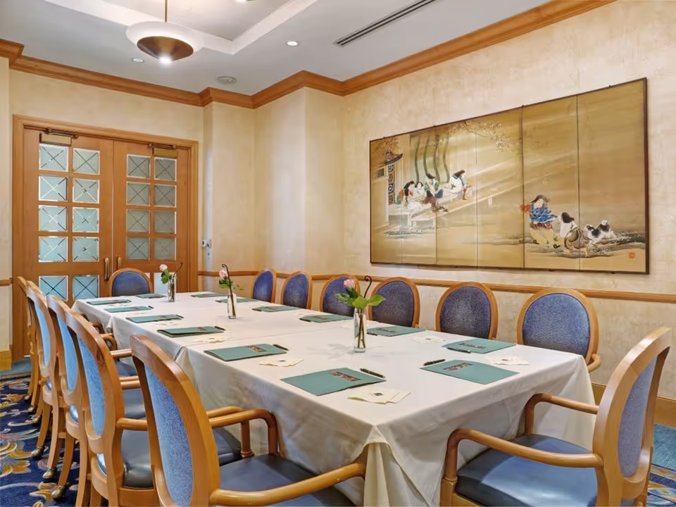A formal dining room with a long rectangular table covered with a white tablecloth. The table is set with green placemats and small vases holding single pink flowers. The room has wooden framed chairs with blue upholstery, beige walls with wood trim, a decorative ceiling light, and a large framed artwork depicting a traditional scene on the wall.