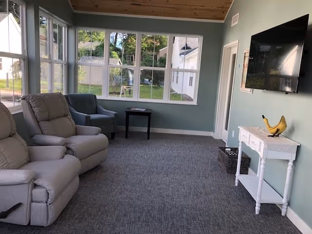 A cozy living room with two beige recliners and a gray armchair arranged near large windows that let in natural light. A small black side table is positioned between the armchair and the windows. On the opposite wall, a flat-screen TV is mounted above a white console table with a decorative yellow banana sculpture. The room has light green walls, a carpeted floor, and a wooden ceiling.