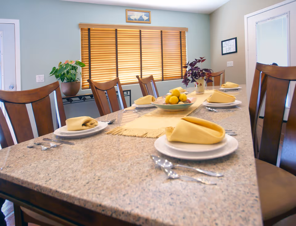 A dining table set with six place settings, each with a white plate, yellow folded napkin, and silverware. The table has a beige runner and a centerpiece bowl with lemons and other fruit. The room has wooden chairs, a window with wooden blinds, a potted plant on a stand, and framed pictures on the walls.