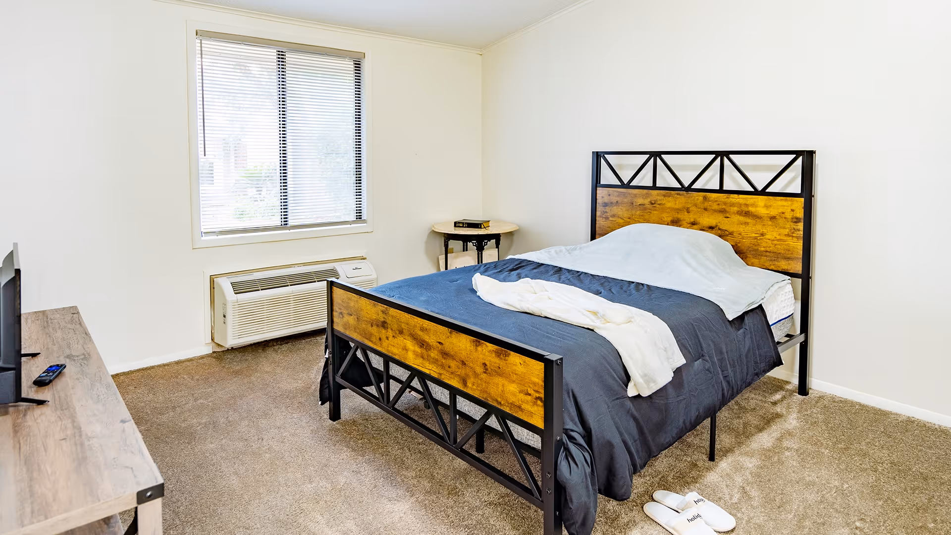 A simple bedroom with a bed featuring a wooden and black metal frame, covered with a blue blanket and a white sheet. There is a small round side table with a book on it next to the bed. A window with blinds is on the wall above an air conditioning unit. A wooden dresser with a TV and remote control is visible on the left side. The floor is carpeted in beige.