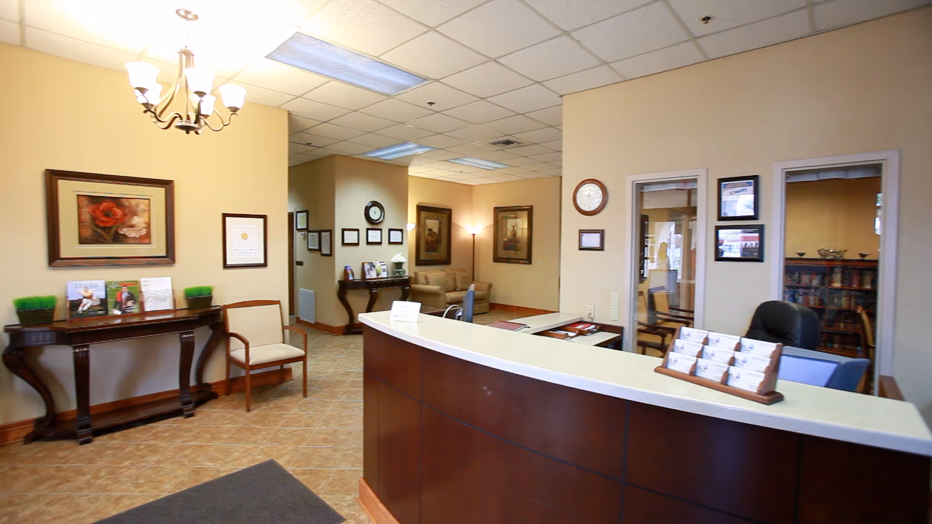 Reception area of a senior living facility with a curved wooden front desk, office chairs, a small seating area with couches, framed artwork and certificates on beige walls, and a chandelier overhead.