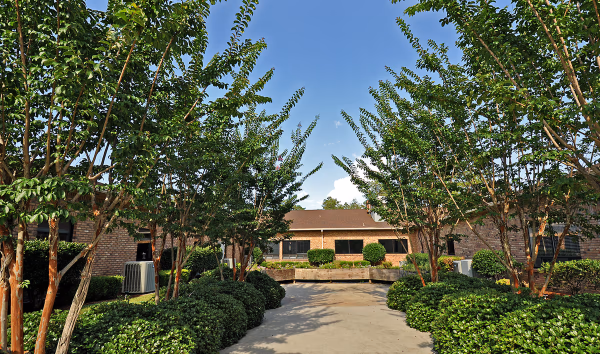 Outdoor courtyard area at Stone County Rehabilitation & Nursing Center with a paved walkway lined by neatly trimmed bushes and tall trees, surrounded by brick buildings under a clear blue sky.