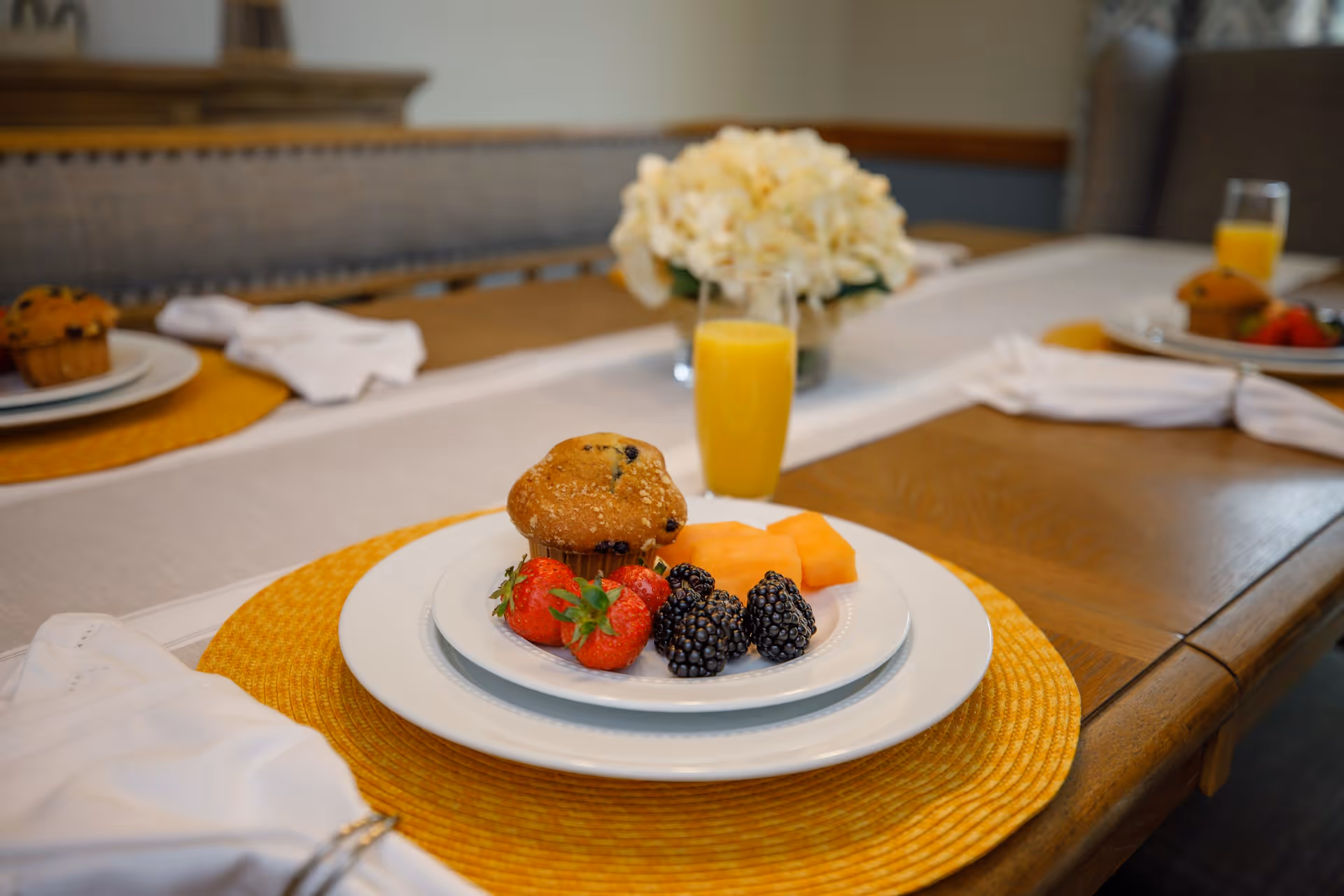 A dining table set with plates containing a blueberry muffin, strawberries, blackberries, and cantaloupe slices, accompanied by a glass of orange juice. The table has yellow placemats and white napkins, with a floral centerpiece in the background.