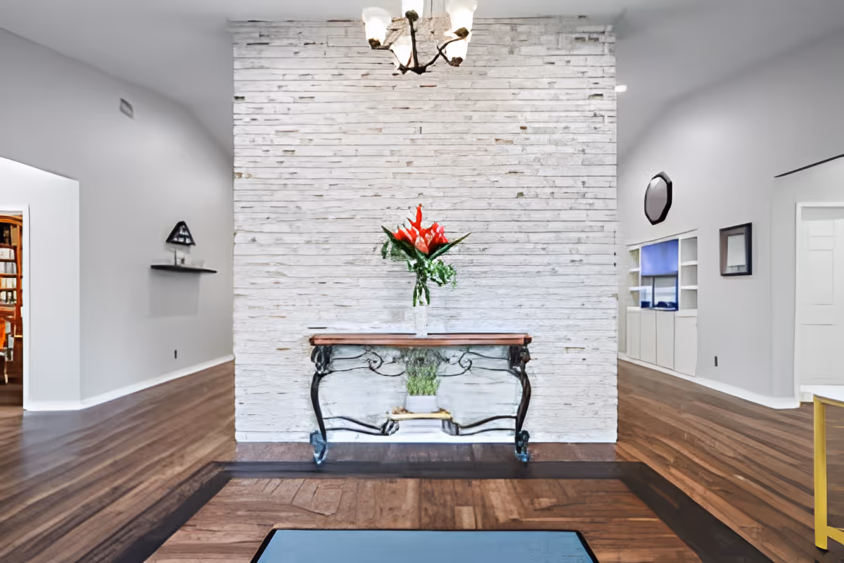 Interior view of a senior living facility hallway with a decorative table holding a vase of red flowers against a white brick wall. The floor is wooden with a patterned inlay, and there is a chandelier hanging from the ceiling. Shelving and framed artwork are visible on the walls in the background.
