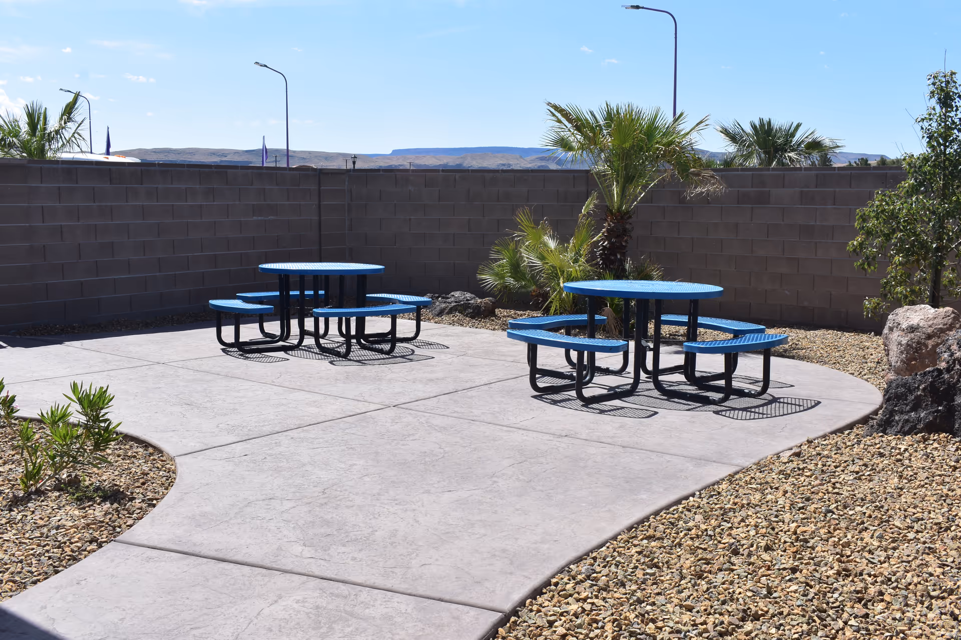 Outdoor patio with blue round picnic tables on a concrete area surrounded by gravel, palm trees, and a cinderblock wall.