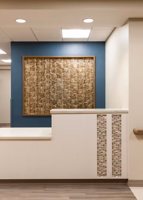 Reception desk area with a white counter and decorative mosaic tile accents on the front. Behind the desk is a blue accent wall featuring a large framed textured artwork with circular patterns. The ceiling has recessed lighting and a white drop ceiling.