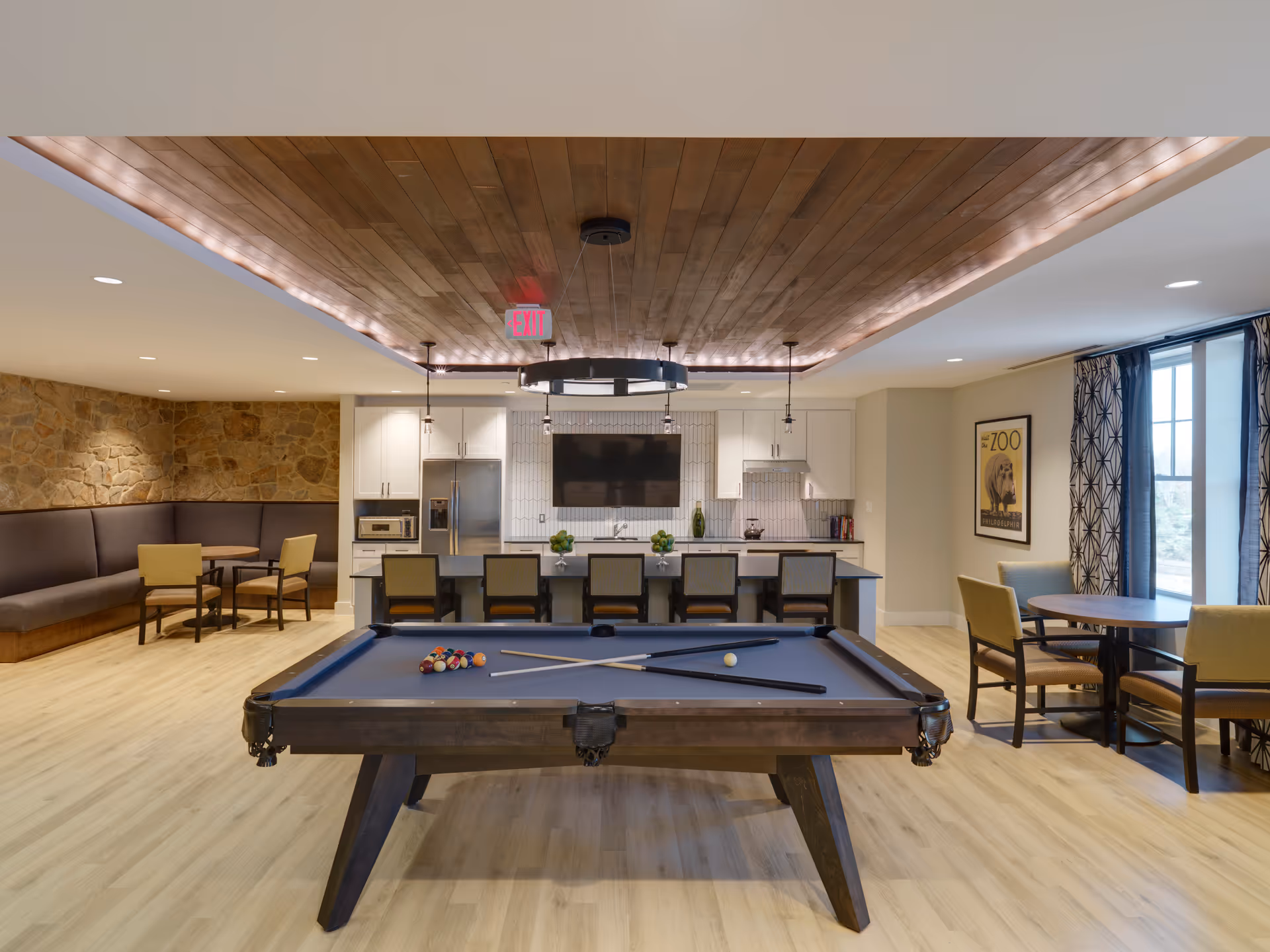 A recreational room featuring a pool table with cues and balls in the foreground, a kitchen area with white cabinets, a refrigerator, and a large TV mounted on the wall in the background. There are bar stools at the kitchen island, a stone accent wall with a curved bench and table seating on the left, and additional table and chairs near a window with patterned curtains on the right. The ceiling has wood paneling with recessed lighting and a circular light fixture above the pool table.