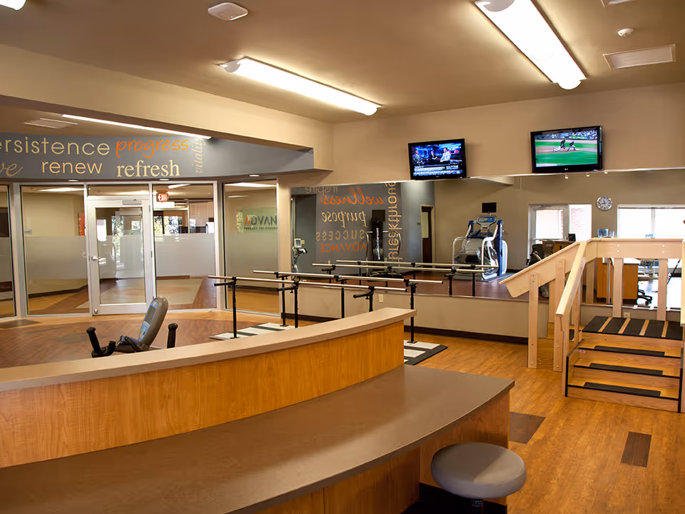 Interior view of a rehabilitation or physical therapy room with wooden flooring, a curved reception desk, exercise equipment, parallel bars for walking practice, a small wooden staircase with handrails, two wall-mounted TVs showing sports and news, and motivational words on the walls.