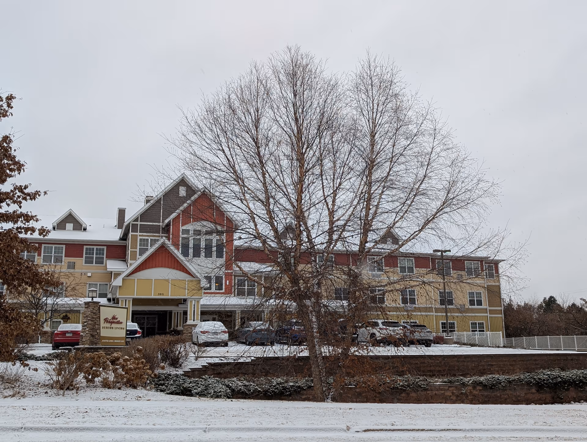 Exterior view of New Perspective Senior Living building in Columbia Heights during winter with snow on the ground and bare trees in front. The building has a multi-colored facade with red, yellow, and beige sections, multiple windows, and a covered entrance. Several cars are parked in front of the building.