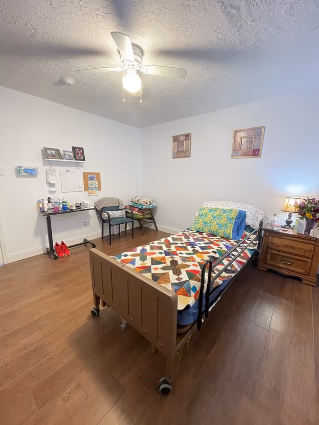 A senior living bedroom with a single hospital-style bed covered with a colorful patchwork quilt. The room has wooden flooring, a ceiling fan with a light, two framed pictures on the wall, a wooden nightstand with a lamp and flowers, a small table with various items, and two chairs against the wall.