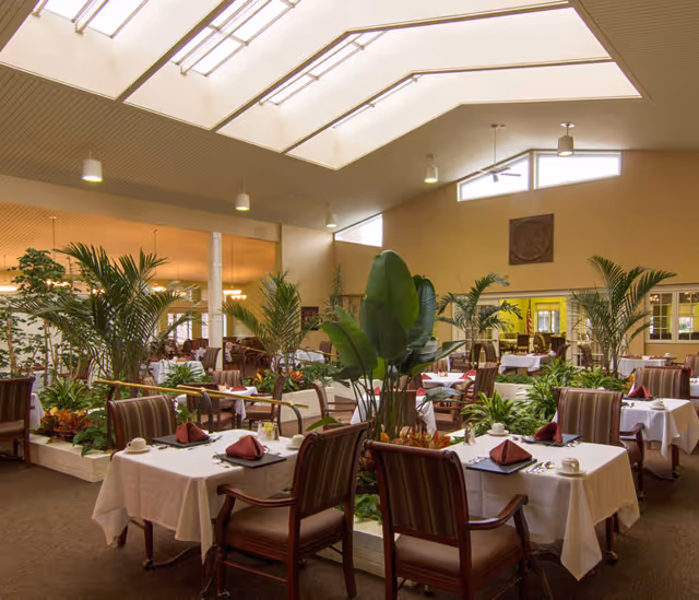 A spacious dining room with multiple tables covered in white tablecloths, each set with maroon napkins, cups, and silverware. The room features large skylights and windows allowing natural light to fill the space. There are several large green plants and planters placed throughout the room, creating a fresh and inviting atmosphere. The chairs are wooden with cushioned seats and backs.