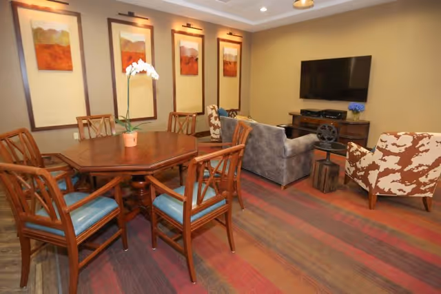 A cozy interior room featuring a wooden dining table with six chairs, a small potted orchid on the table, a seating area with two patterned armchairs and a gray sofa facing a wall-mounted flat-screen TV, and four framed landscape paintings on the wall.