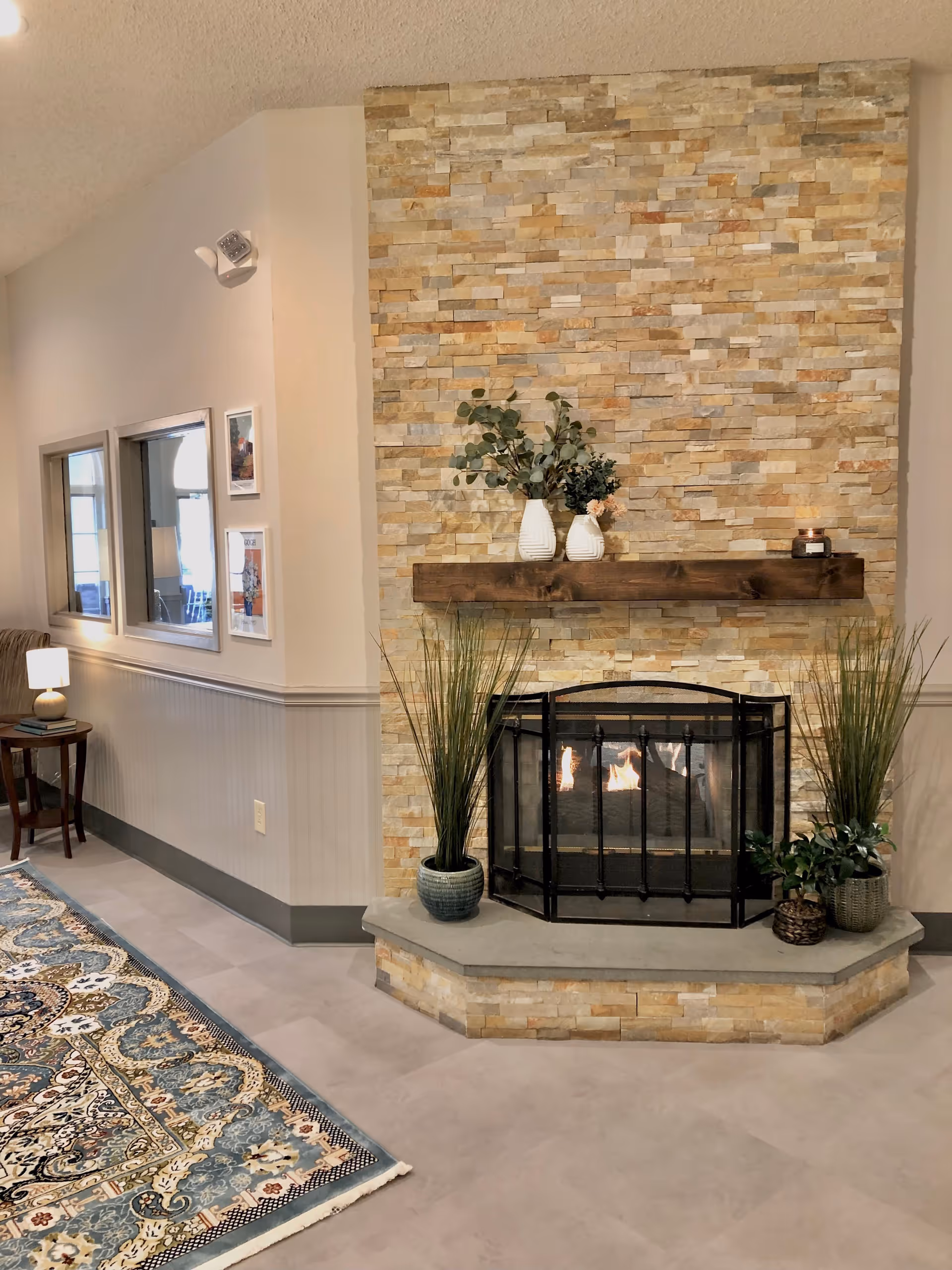 A cozy indoor living area featuring a stone fireplace with a wooden mantel. On the mantel are two white vases with green and pink floral arrangements and a small candle. The fireplace has a black metal screen in front of it. On the hearth, there are three potted plants with tall green grass and leafy foliage. To the left, there is a small wooden side table with a lamp and some books, next to a patterned area rug with blue, beige, and black designs. The walls are light-colored with a chair rail and wainscoting, and there are two windows and framed pictures on the wall.