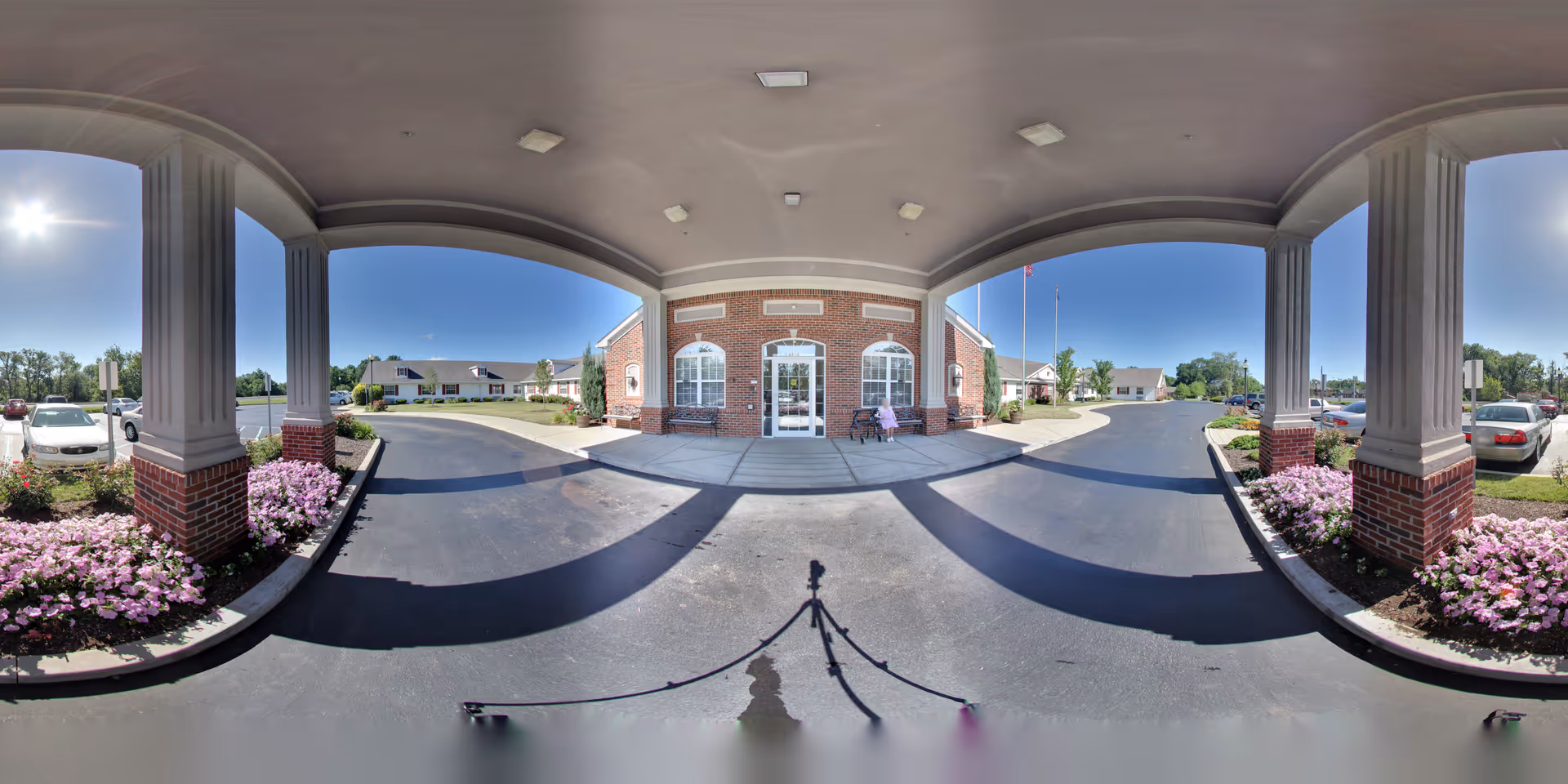 Covered porte-cochère and main entrance of a brick senior living facility with columns, flower beds and parked cars.