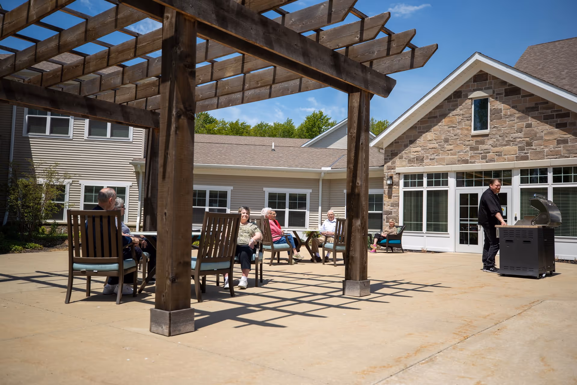 Older adults sit on a patio under a wooden pergola at a senior living facility while a person stands at a grill near the building.