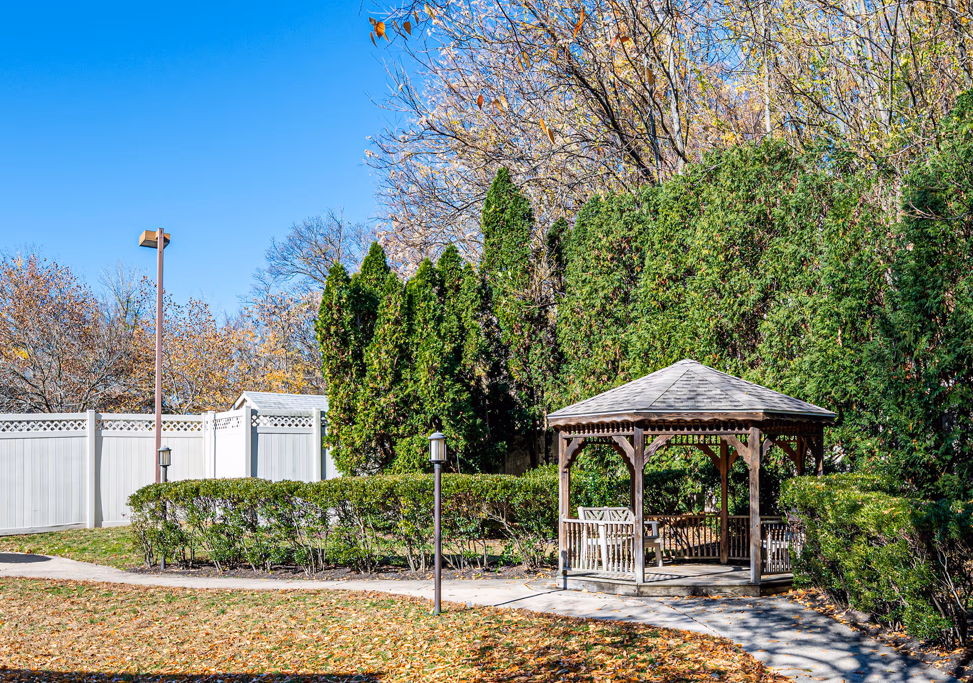 Outdoor garden area with a wooden gazebo surrounded by green bushes and tall trees. A paved walkway leads to the gazebo, and a white fence encloses the area under a clear blue sky.