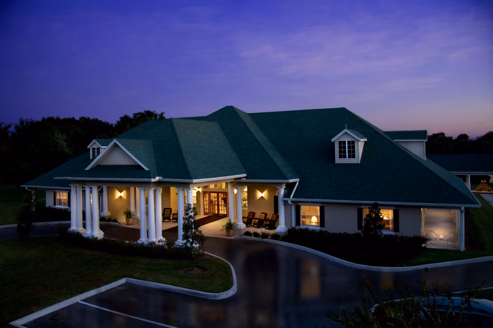 Front entrance of a single-story senior living building with white columns and a green roof lit at dusk.