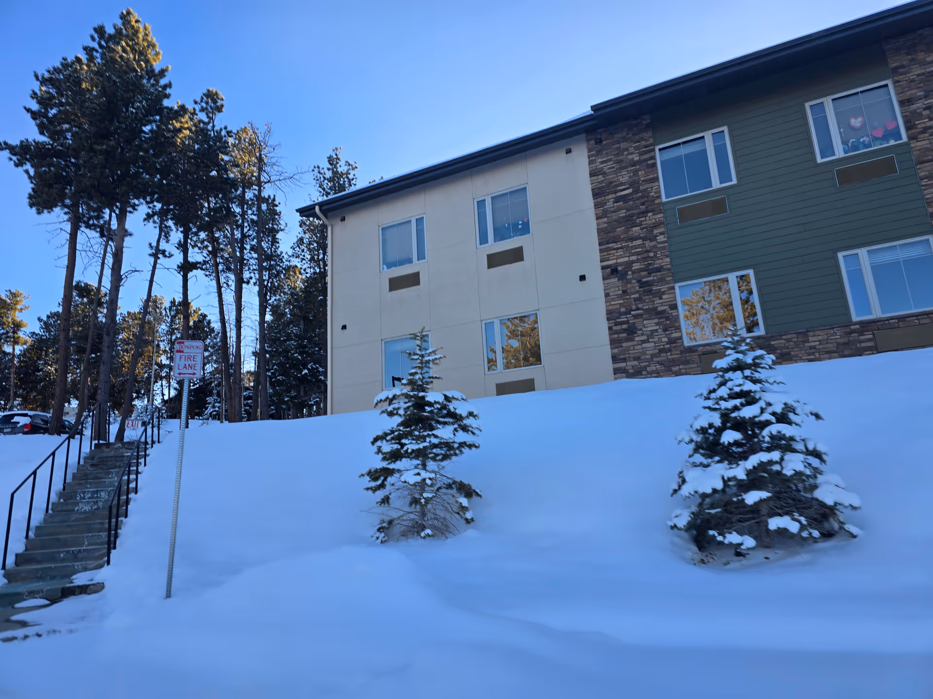 Snow-covered front of a multi-story senior living building with small evergreen trees, a stairway, and a 'Fire Lane' sign under a clear blue sky.