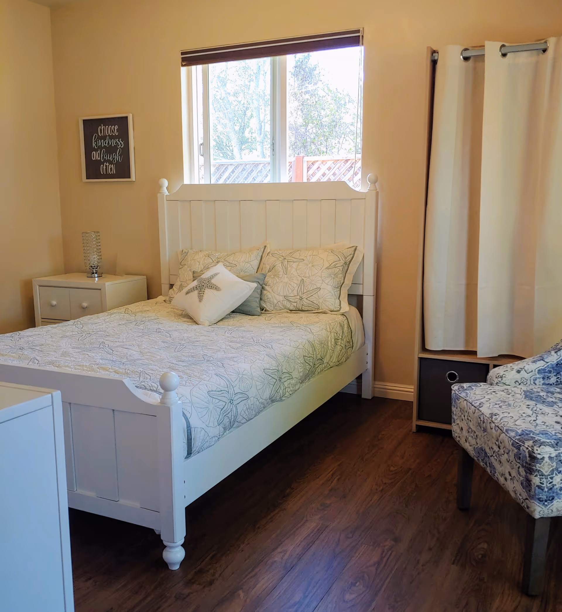 A bright bedroom with a white wooden bed, patterned bedding, a nightstand, window, and an upholstered chair.