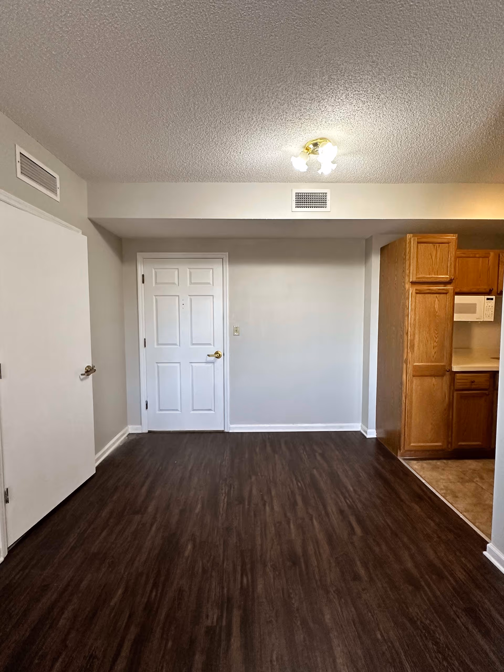 Empty interior room with dark wood flooring, white walls, a white door in the center, and a kitchen area with wooden cabinets and a microwave on the right side.