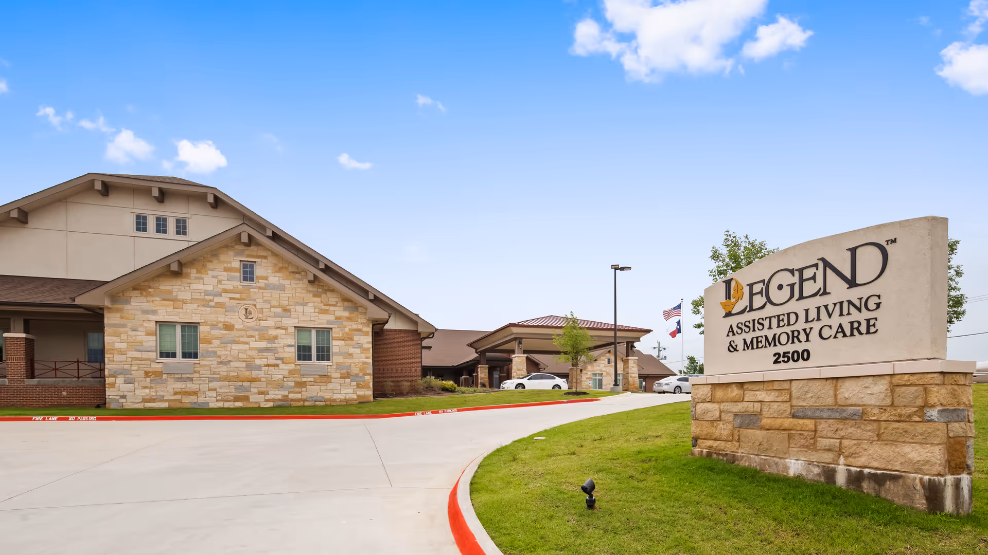 Exterior view of Legend Assisted Living & Memory Care facility showing a stone and brick building with a driveway and a large sign displaying the facility name and address 2500. The sky is clear with a few clouds.